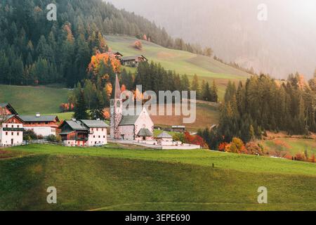 Pittoresco paesaggio alpino della storica chiesa in pietra di Santa Maddalena su una lussureggiante collina verde tra pini autunnali e vette delle Odle Dolomites Foto Stock