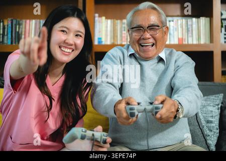 Giovane donna in punti rosa felicemente. Un uomo più anziano sorride, ha i controller di gioco. Condividono un comodo divano in una camera piena di libri. Gioiosa intergeneraziona Foto Stock