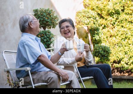 Una coppia di anziani siede su sedie bianche da giardino. Tengono tazze da tè bianche, ridendo a vicenda. I cespugli e l'erba verdi lussureggianti li circondano. La loro affectiona Foto Stock