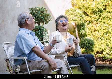Le coppie di anziani si rilassano sulle sedie da giardino. La donna ride, tenendo in mano una tazza da tè. L'uomo sorride, si gode la compagnia. Lussureggianti arbusti e alberi verdi li circondano. Soleggiato Foto Stock