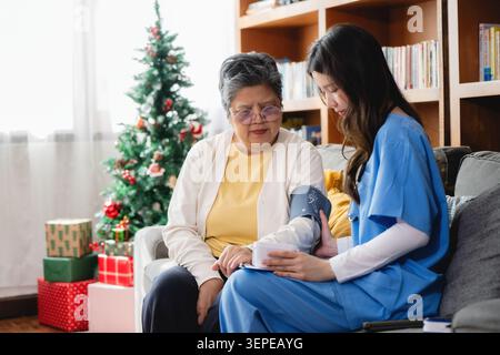 Infermiera in scrub blu controlla la pressione sanguigna su una donna anziana espressione preoccupata si siede sul divano. Mostra sullo sfondo l'albero di Natale decorato e il regalo Foto Stock