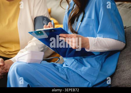 Infermiere in scrub blu controlla la pressione sanguigna, l'operatore sanitario tiene gli appunti mentre misura la pressione sanguigna con il bracciale sul paziente. Il paziente si siede comodamente Foto Stock