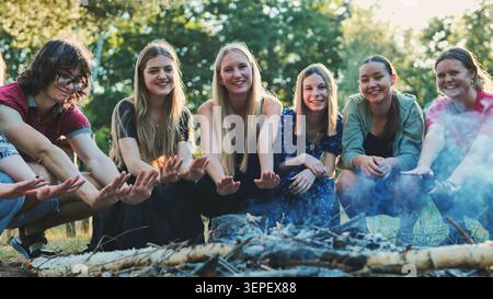 Gli studenti si accoccolano intorno al fuoco scoppiettante, condividendo calore e cameratismo durante la riunione serale estiva nei boschi Foto Stock