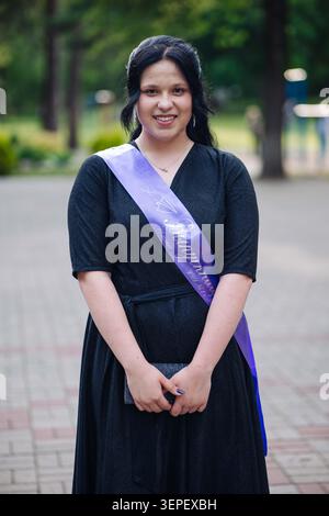 Giovane studente laureato che indossa una fascia di laurea, sorridendo brillantemente mentre festeggia durante una cerimonia di laurea all'aperto nel parco Foto Stock