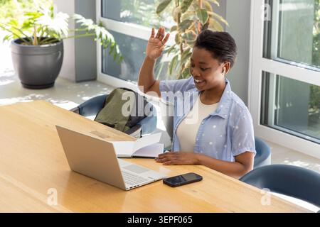 Donna afro-americana seduta e salutando mentre studiava nella sala comune con laptop e notebook Foto Stock