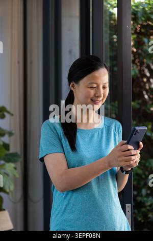Donna asiatica che indossa T-shirt e smartwatch blu, tiene il telefono scuro e sorride da una porta scorrevole in vetro Foto Stock