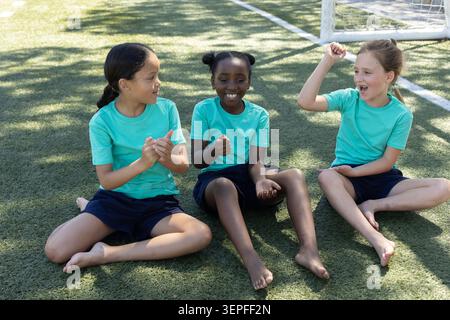 Tre ragazze in età scolare sedute su un tappeto erboso vicino a rete porta con magliette color ottanio pantaloncini navy, applausi Foto Stock