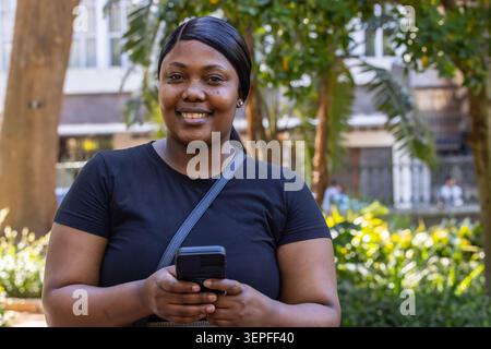 Donna africana adulta in piedi nel cortile con alberi in una maglietta nera che regge il telefono mostrando la cinghia della borsa Foto Stock