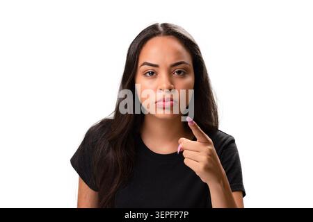 Giovane donna ispanica seria e concentrata che guarda direttamente la telecamera mentre punta il dito indice in avanti. Foto in studio che rappresenta la scelta, accu Foto Stock