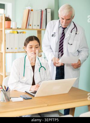 Medico anziano esperto e giovane infermiera donna che lavorano insieme con case history su laptop al tavolo dell'ufficio della clinica Foto Stock