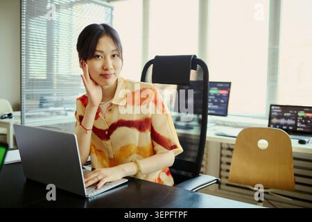 Ritratto di una giovane assistente d'affari asiatica o segretaria che lavora su un laptop in ufficio, sorridendo e guardando la fotocamera. Ideale per la produttività sul luogo di lavoro, le startup e le comunicazioni aziendali Foto Stock