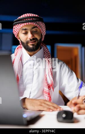 Uomo arabo che lavora in modo efficiente sul posto di lavoro navigando su Internet e scrivendo appunti su un libro; ragazzo musulmano che fa uno studio con il suo blocco appunti e il suo personal computer. Foto Stock