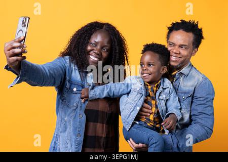 I genitori orgogliosi e felici si divertono a scattare foto con il loro simpatico bambino su sfondo giallo, catturando momenti divertenti insieme, con una felice coppia afroamericana che posa per i selfie con il figlio. Foto Stock