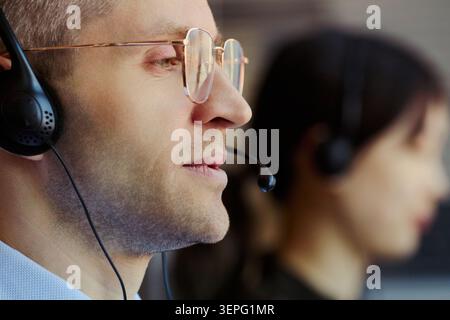 Primo piano di un giovane uomo caucasico che indossa le cuffie, assistendo i clienti nel call center mentre una collega lavora nelle vicinanze. Ideale per assistenza clienti, CRM, outsourcing Foto Stock