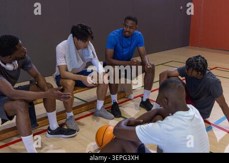 Diversi giocatori di basket maschili in abbigliamento sportivo seduti sulla panchina e sul campo in palestra, che tengono il basket Foto Stock