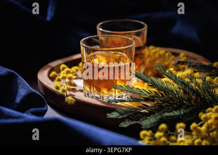 Bicchieri da whisky o Cognac con fiori di Mimosa su vassoio di legno scuro elegante drink Foto Stock