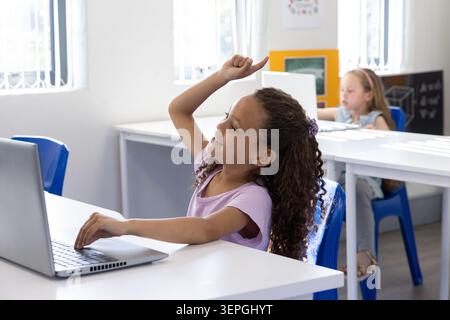 Ragazze in classe sedute in classe alle scrivanie, alzando la mano davanti mentre si utilizza un computer portatile color argento Foto Stock