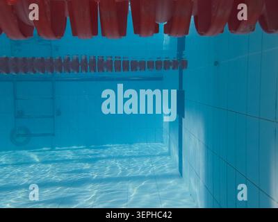Vista subacquea della piscina con divisorio galleggiante rosso in primo piano, scala per piscina, piastrelle blu e riflessi solari. Foto Stock