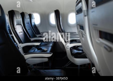 File di sedili aerei non occupati con cinture di sicurezza, poggiatesta e tavolini visibili all'interno di una moderna cabina per aeromobili commerciali a fusoliera stretta Foto Stock
