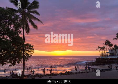 Vibrante tramonto su Magic Sands Beach sulla Big Island delle Hawaii, con silhouette di palme, onde dell'Oceano Pacifico e bagnanti che godono della serata tropicale. Foto Stock