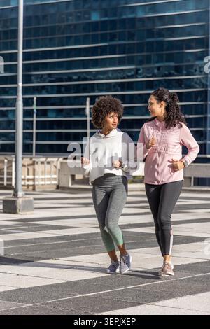 Diverse amiche che camminano attraverso la piazza a strisce, attraverso un edificio di vetro, indossando scarpe da running. Urban, outdoor, duo, sneakers, athleisure, allenamento, luce del sole Foto Stock