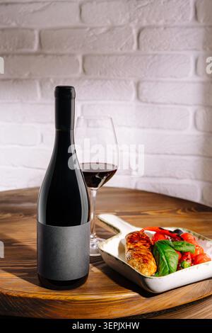 Una bottiglia di vino rosso si trova accanto a un bicchiere pieno di vino. Il pollo alla griglia con pomodori e verdure fresche è su un piatto. L'ambiente e' semplice, con un tavolo in legno e pareti in mattoni bianchi. Foto Stock