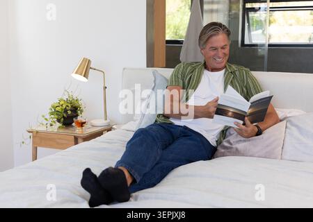 Uomo maturo sdraiato sul letto indossando un orologio da polso che legge un libro cartaceo aperto a casa con tè. Uomo, camera da letto, accogliente, naturale, luce del sole, verde, legno Foto Stock