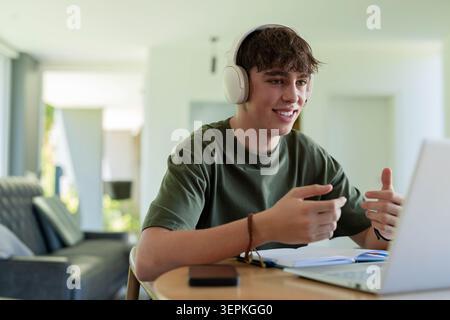 Adolescente maschio che indossa una cuffia bianca che passa al laptop argentato su un tavolo rotondo con notebook in casa Foto Stock