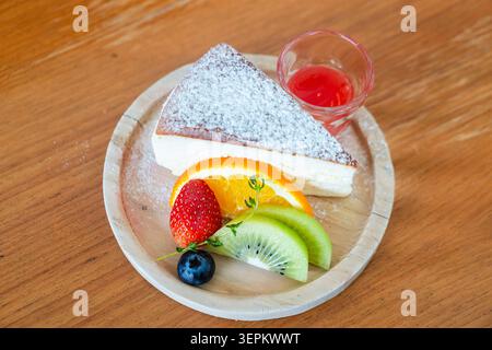 Una fetta di cheesecake cremosa ricoperta di zucchero a velo, servita con fette fresche di fragola, kiwi, arancia e un bicchiere di succo di frutta su una Foto Stock