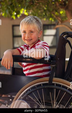 Bambino seduto in sedia a rotelle nel cortile, con tee a righe rosse bianche e sorridente Foto Stock