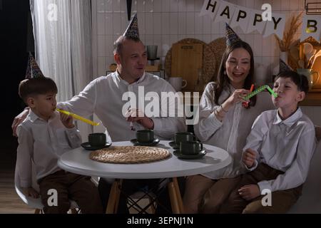 Buon compleanno famiglia caucasica intorno alla torta, genitori e due ragazzi che indossano cappelli da festa, soffiatori, bottiglie di champagne sul tavolo, banner e palloncini Foto Stock