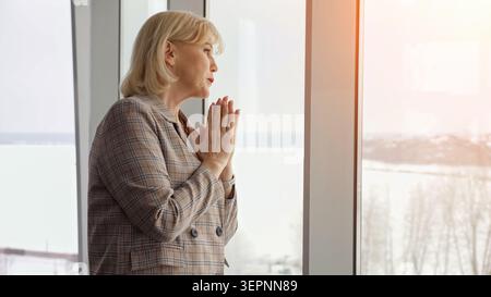 Donna d'affari anziana con le mani strette che guarda fuori da una grande finestra mentre contempla, riflette e si sente preoccupata, rappresentando lo stress Foto Stock