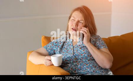Donna bianca sorridente sul divano arancione che immerge la luce del sole calda mentre parla al telefono tenendo in mano un abito floreale da tazza da caffè rilassato e gioioso Foto Stock