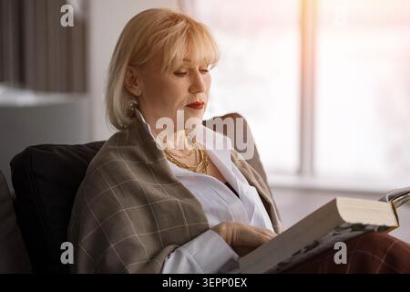 Donna anziana che si rilassa sul divano di casa, si concentra su un libro durante il tempo libero, godendo un momento tranquillo di invecchiamento in comfort e pensione Foto Stock