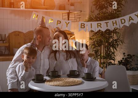 Festa di compleanno in famiglia al tavolo da cucina, calda scena serale con genitori e bambini che indossano cappelli da festa, banner di buon compleanno sullo sfondo Foto Stock