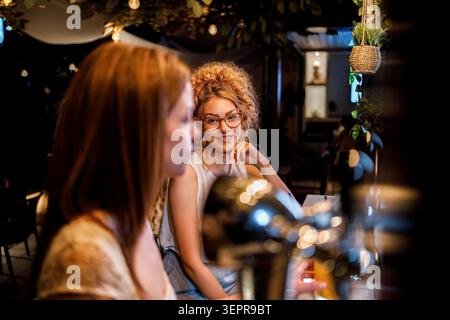Due amici si siedono a un tavolo da caffè la sera. Un'amica sorride mentre l'altra la guarda verso di lei. Si divertono insieme. Foto Stock