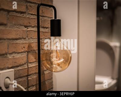 Una lampadina a incandescenza d'epoca è appesa a una parete nera accanto a una parete in mattoni a vista, con un globo di vetro ambrato, filamento visibile, calda luce soffusa Foto Stock