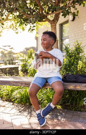 Bambino afroamericano seduto sulla panchina nel cortile tenendo un tablet e sorridendo accanto allo zaino Foto Stock