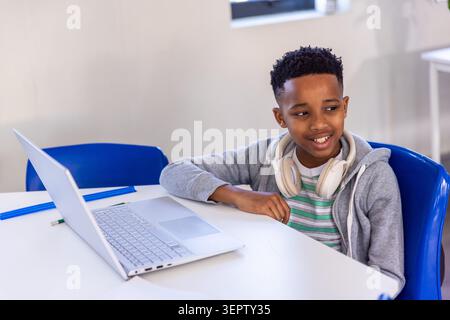 Bambino afroamericano seduto in classe con felpa con cappuccio grigia, cuffie bianche, laptop argento Foto Stock