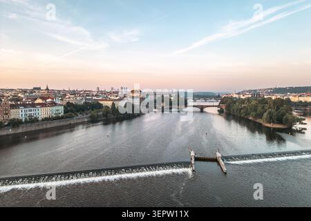 Una vista della diga sul fiume Moldava a Praga. L'acqua scorre sopra la struttura mentre la città è illuminata dal tramonto. Ponti e alberi fiancheggiano le rive. Foto Stock