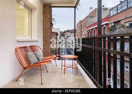 Una piccola terrazza al quinto piano con vista sui tetti, arredata con una panca in vimini, cuscini colorati e una tenda in tela beige che filtra il sole Foto Stock