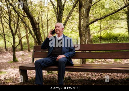 Uomo anziano seduto su una panchina del parco che parla con un cellulare Foto Stock