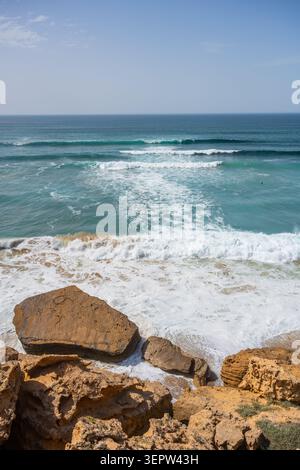 Le onde si infrangono sulla costa rocciosa mentre l'Oceano Atlantico incontra la terra nel sud del Marocco. Il cielo è limpido e l'acqua è una vibrante tonalità di blu sotto la luce del sole. Foto Stock
