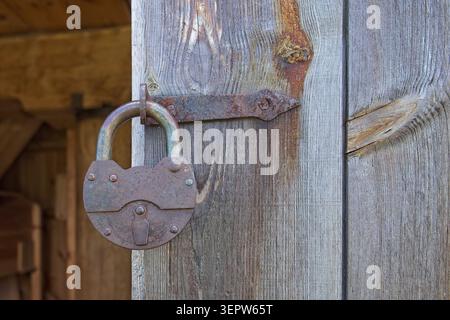 Primo piano di un lucchetto vintage in ghisa fortemente arrugginito che fissa una porta in legno tramite un'antica cerniera in metallo, Estonia, Europa. Foto Stock
