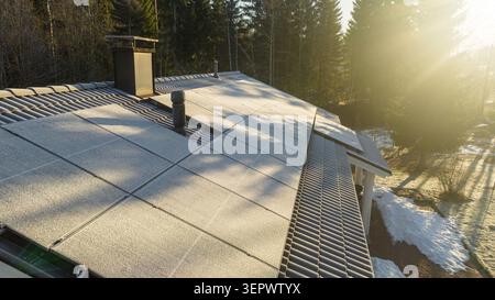 Vista aerea dei pannelli solari ricoperti di ghiaccio su un tetto di una casa piastrellata con una foresta sempreverde e la luce solare invernale a Uusimaa, Finlandia. Foto Stock