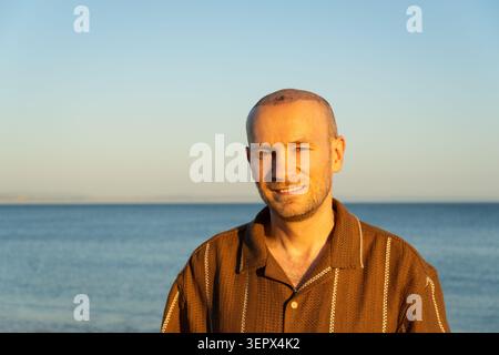 Confident Smiling Man in Brown Polo with Hair Transplant Against Sea Foto Stock