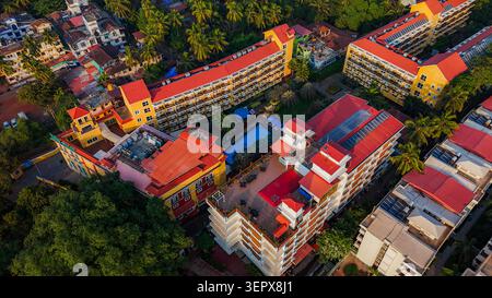 Vista aerea di un vivace complesso turistico con tetti di tegole rosse e una piscina centrale circondata da lussureggianti palme a Calangute, Goa, India. Foto Stock