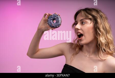 Una giovane donna si aspetta di mangiare ciambella smaltata viola con spruzzi su un vivace sfondo rosa Foto Stock