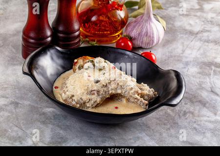 Cosce di pollo arrosto in salsa cremosa con capperi e grani di pepe servite in una ciotola nera, circondate da aglio, pomodori, olio d'oliva e peperoni su S Foto Stock