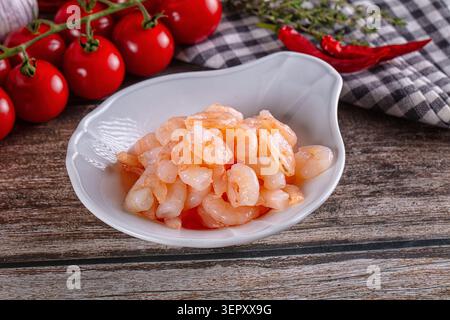 Mucchio di gamberi pelati cotti in un recipiente di ceramica bianca su una superficie rustica in legno. Pomodori freschi, peperoncino rosso, timo e aglio sullo sfondo. H Foto Stock